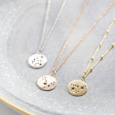 silver constellation disc charm on silver belcher chain necklace laying next to rose gold pisces constellation disc charm on rose gold belcher chain necklace and gold constellation disc charm on gold bead chain necklace